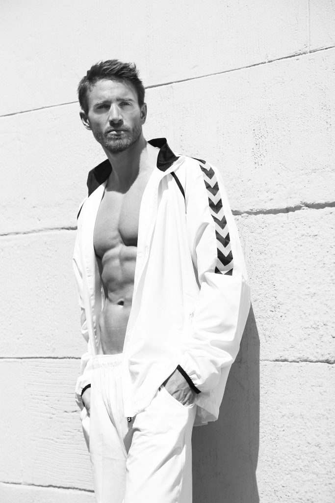 Photo of an athletic man in a white tracksuit leaning against a facade of large stone blocks.