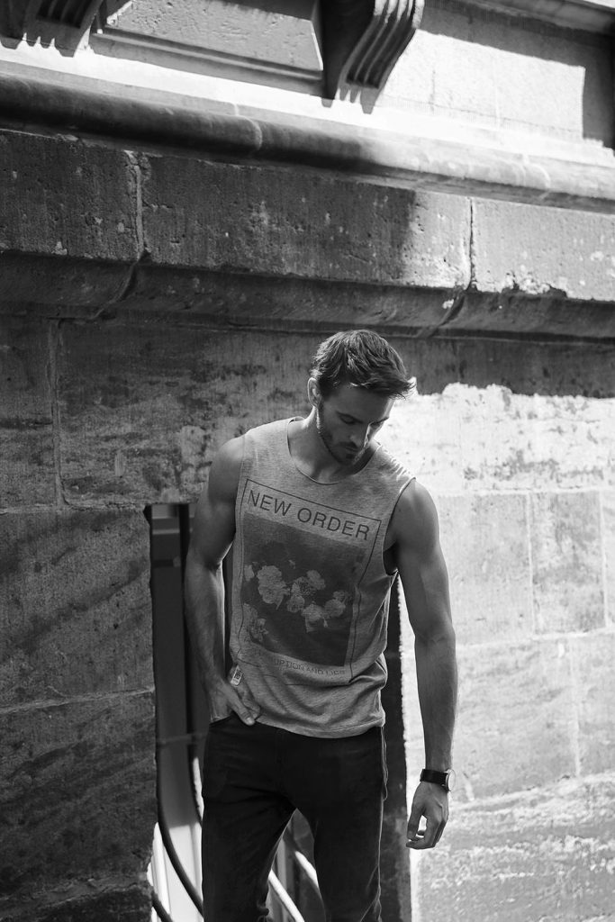 Black-and-white portrait of bearded male model in tank-top standing in front of a stone wall.