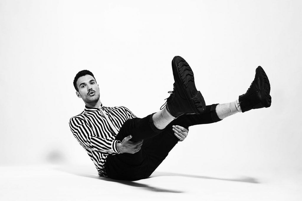 Black-and-white photo of male model wearing vintage striped shirt and sitting on the floor with his legs up in the air.